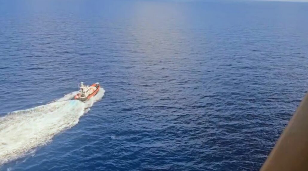 A small red and white boat, using its propulsion system, travels across calm, deep blue Mediterranean waters, leaving a visible wake behind it. The photo is taken from an elevated perspective, possibly from a mega cruise ship.