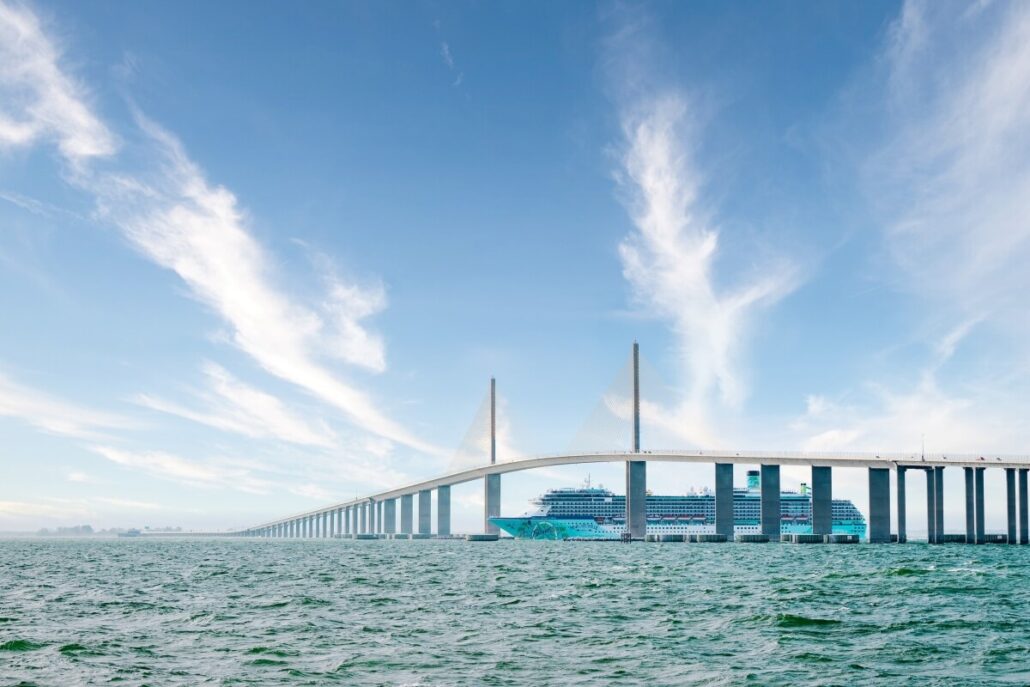 A large cruise ship from a top cruise line passes under a long cable-stayed bridge spanning across a body of water, with blue sky and streaks of white clouds overhead.