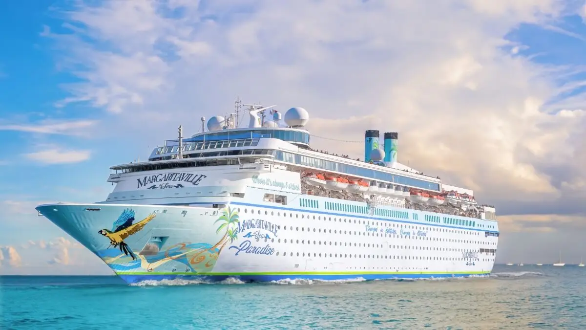 A large white cruise ship labeled "Margaritaville at Sea Paradise" sails on calm blue water under a partly cloudy sky, featuring tropical-themed artwork on its hull and promoted as an all-you-can-sail pass by the cruise line.