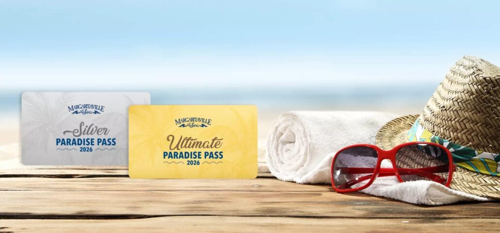 Two cards labeled “Silver Paradise Pass 2026” and “Ultimate Paradise Pass 2026” from a leading cruise line rest on a wooden surface beside a towel, straw hat, and sunglasses, offering an all-you-can-sail pass with the beach in the background.