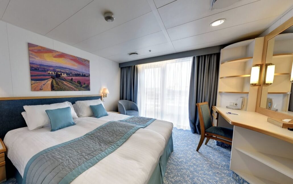 A hotel room with a double bed, blue and white bedding, a painting above the bed, a desk with chair and mirror, shelves, an armchair, and a window letting in natural light—ideal for Villa Vie Residences or Lifetime Living at Sea experiences.