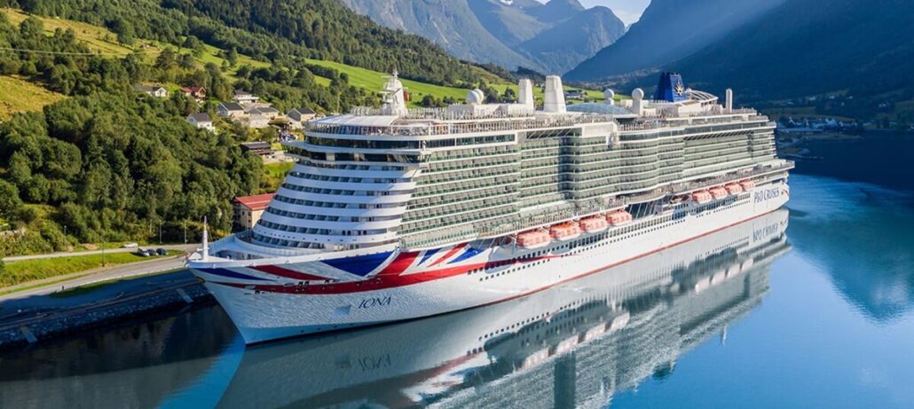 A large white cruise ship with red and blue markings is docked by a calm body of water, surrounded by green hills, scattered houses, and distant mountains under a clear sky after a brief galley fire led to a temporary restaurant closure.