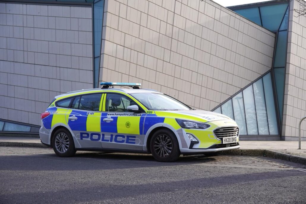 A marked police car is parked on the street in front of a modern building with geometric glass and metal panels, responding to a potential bomb threat involving cruise passengers from a UK ship. The vehicle features blue and yellow police livery.