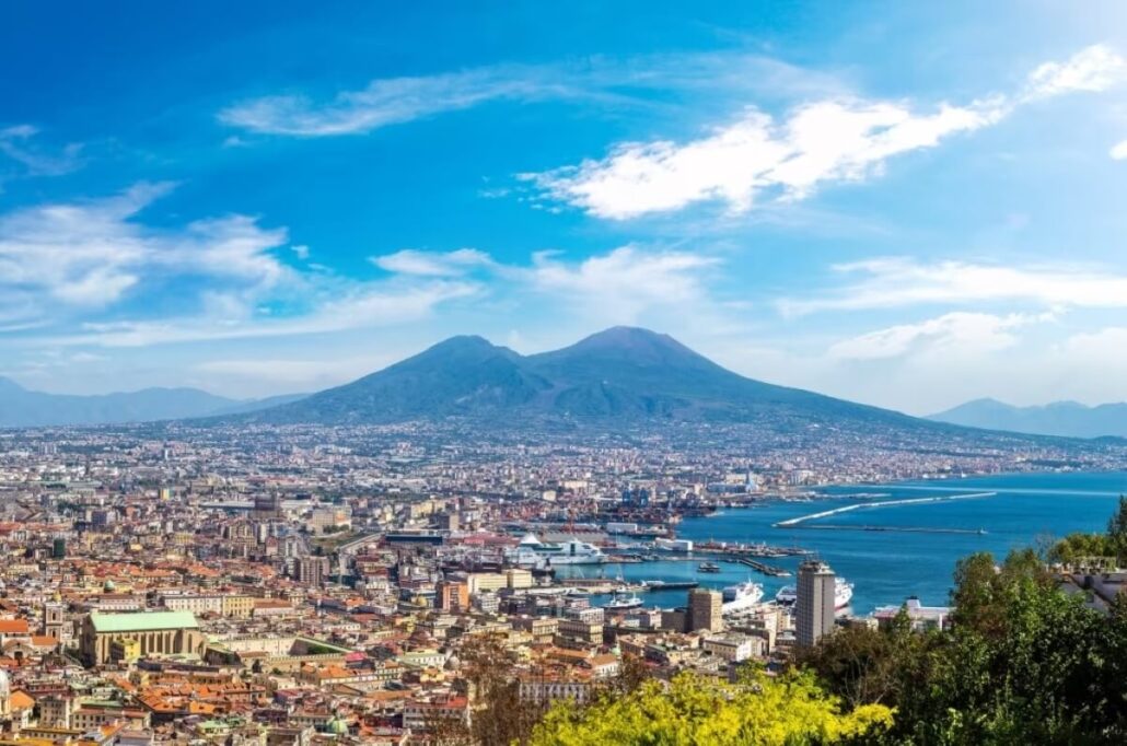 A panoramic view of Naples, Italy, with a dense cityscape in the foreground and a cruise ship gliding past Mount Vesuvius under a blue sky with scattered clouds.