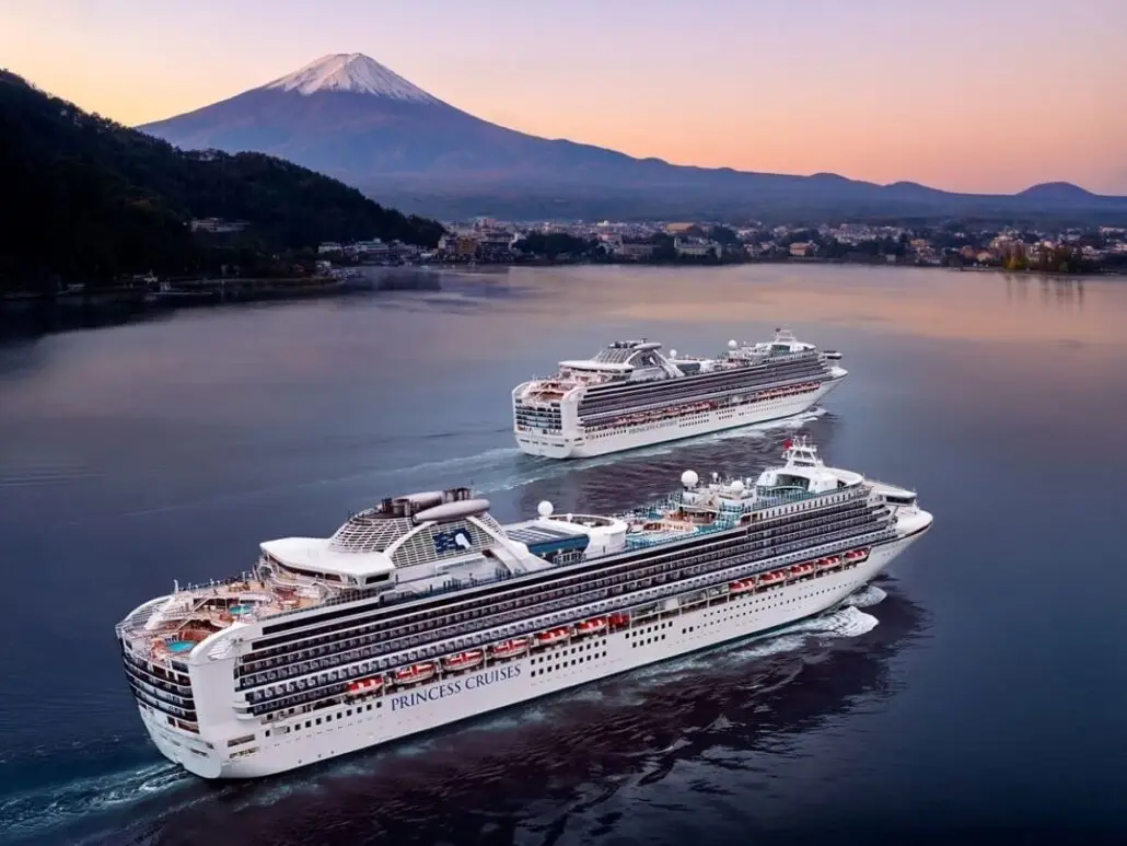 Two Princess Cruises ships, including Royal Princess, sail on calm water near a shoreline dotted with buildings. In the background, Mount Fuji rises under a pastel sky, highlighting the beauty of Asia sailings at sunrise or sunset.