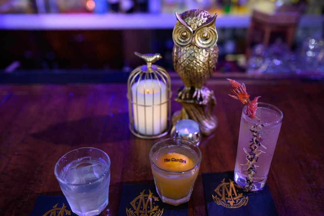 Three cocktails are arranged in a row on a bar with decorative coasters. Behind them, a lit candle in a small birdcage and a gold owl ornament evoke the Spellbound charm of a Magic Castle. The background features blurred bar shelves.
