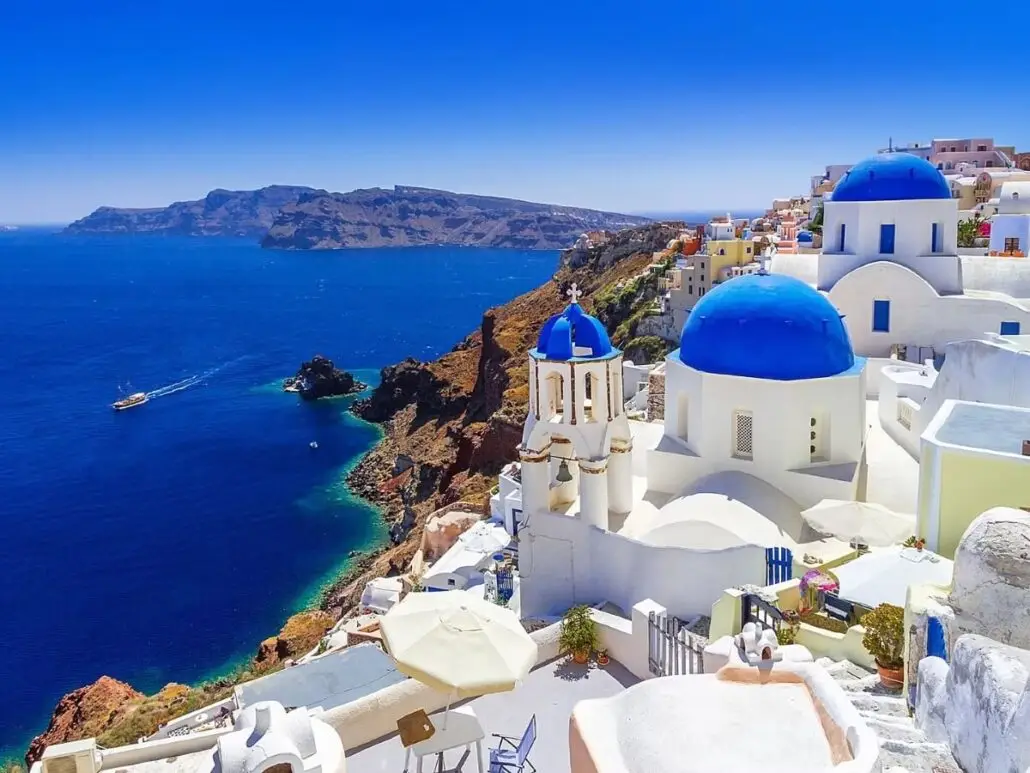 A seaside view of white buildings with blue domes on a cliffside in Santorini, Greece, overlooks deep blue water with a cruise boat and distant hills under a clear blue sky.