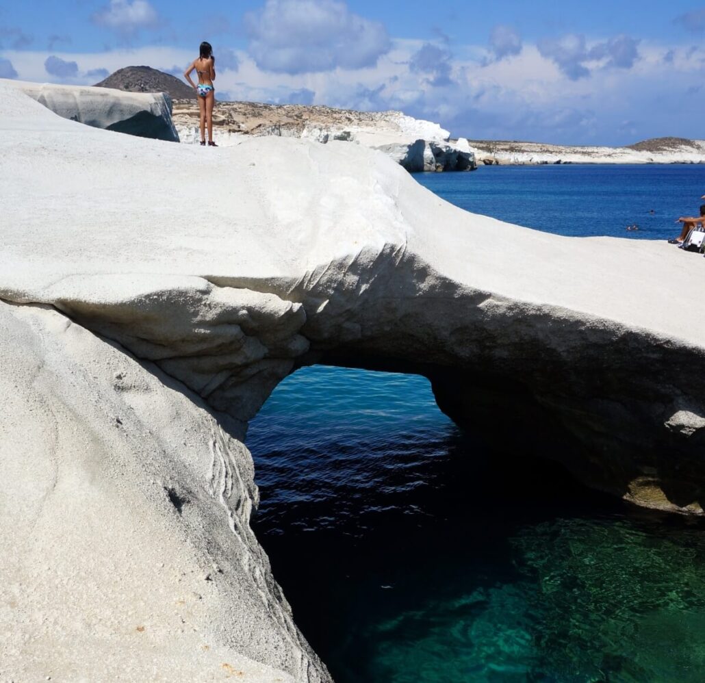 A person in swimwear stands on a white rock formation above a natural stone arch by clear blue water on a stunning Greek island, with a rocky coastline and hills in the background under a partly cloudy sky.