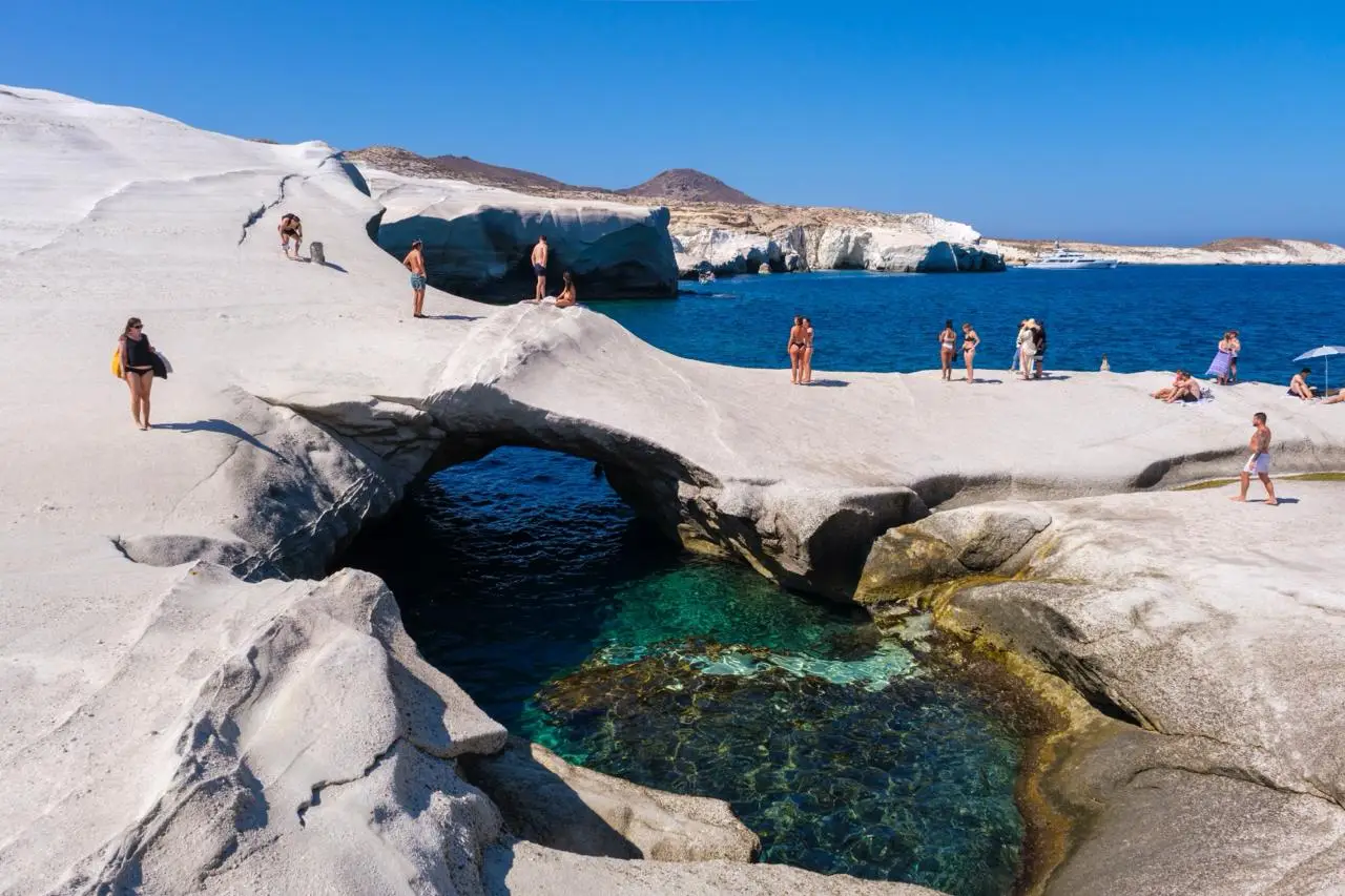 People stand and walk on smooth, white rock formations overlooking clear blue water and a natural arch on a Greek island. Some guests from a cruise ship are near the edge, while others explore the rocky landscape under the sunny sky.