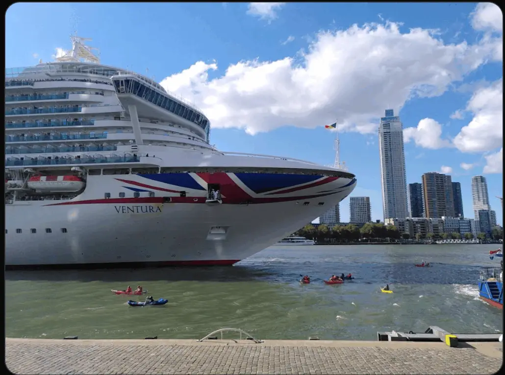 A large cruise ship named Ventura sails near a city waterfront with several kayakers in the water. Modern high-rise buildings and trees are visible in the background under a partly cloudy sky.