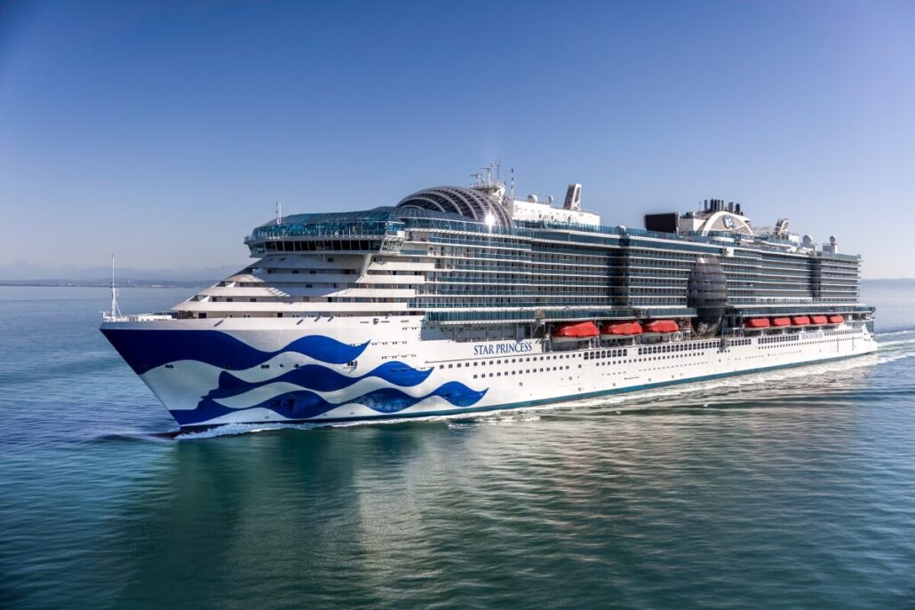 The Star Princess sails on calm, clear water under a blue sky during its sea trials. The large white and blue cruise ship features multiple decks, lifeboats along the sides, and its name proudly displayed on the hull.