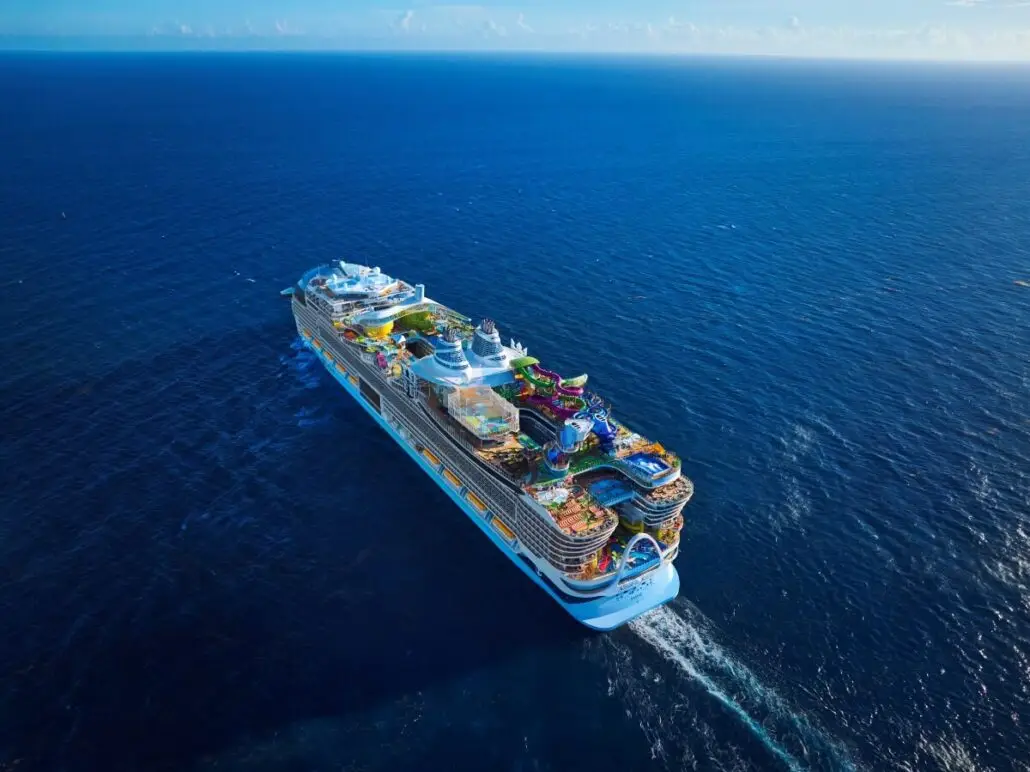A large, colorful Royal Caribbean cruise ship sails on calm blue ocean water under a mostly clear sky, viewed from above and behind. The upper deck features various attractions and amenities, echoing the elegance of Diana Ross in her Godmother Role.