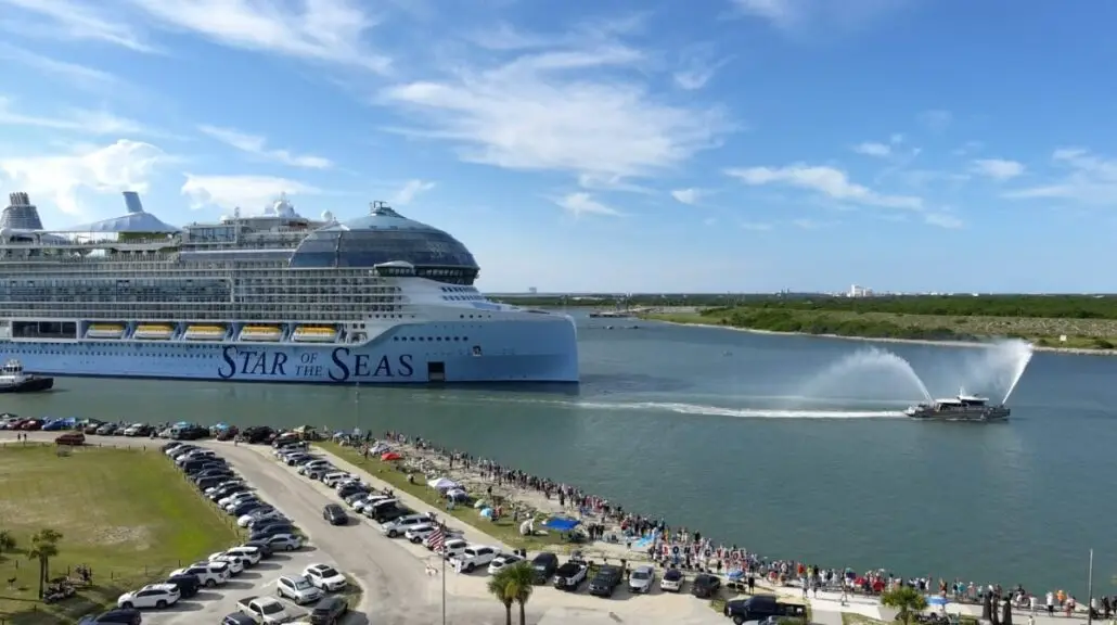 A large cruise ship, "Star of the Seas," makes its inaugural sailing near Port Canaveral’s shoreline as a crowd watches and a smaller boat sprays water jets. Parked cars line the waterfront road beneath a partly cloudy sky.