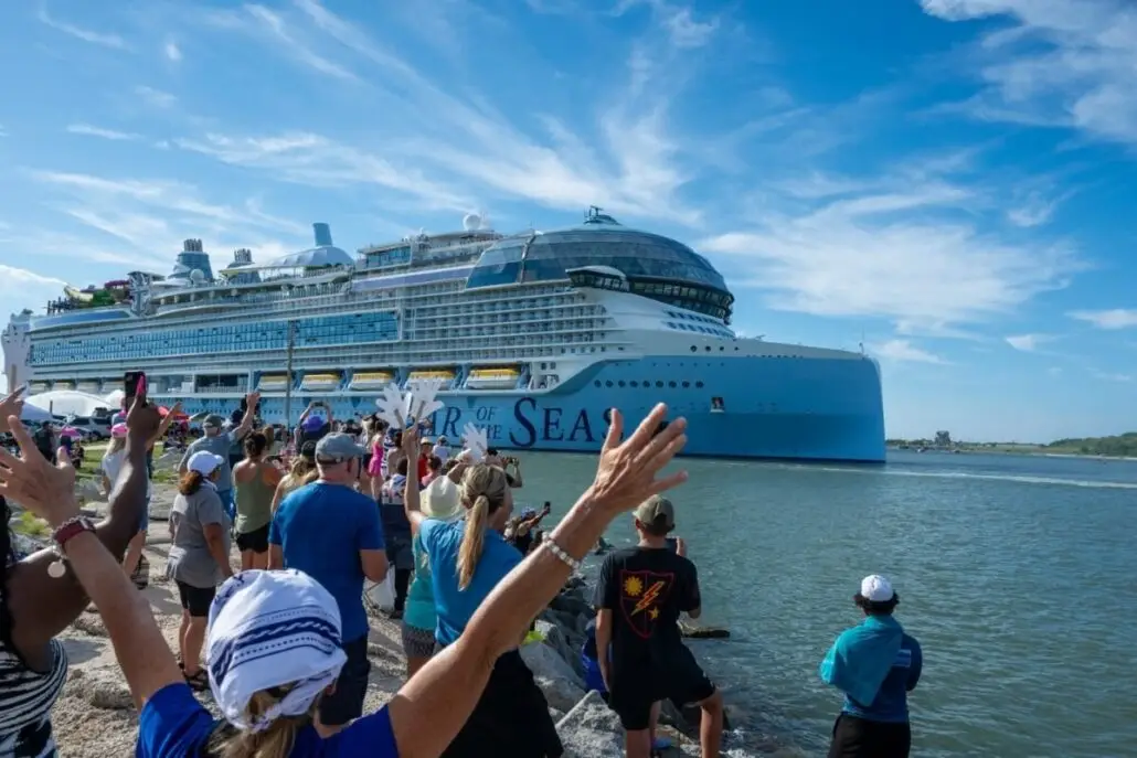A large cruise ship labeled "Oasis of the Seas" sails near the shore from Port Canaveral while a crowd of people, some with hands raised, watch and wave from the waterfront on a sunny day.