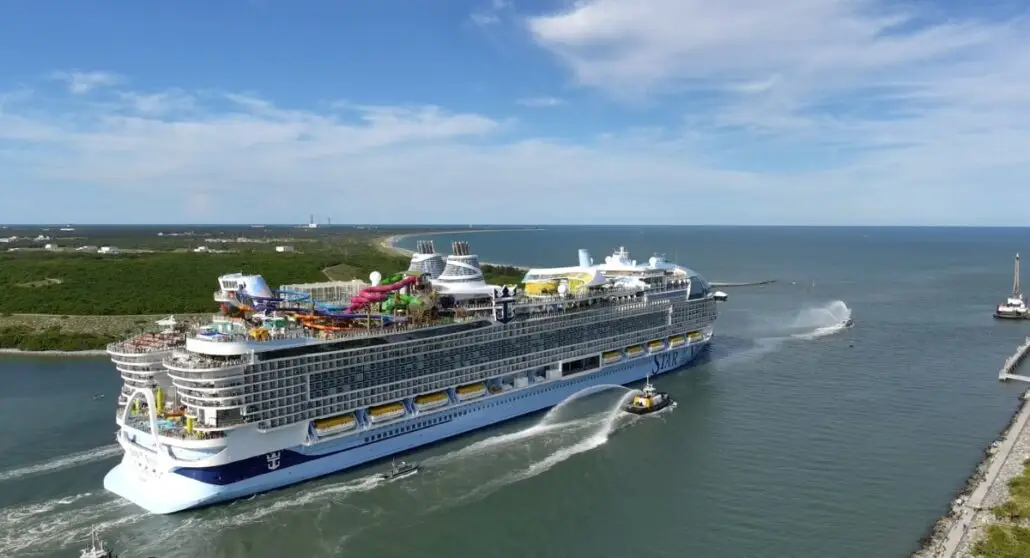The Star of the Seas, on its inaugural sailing from Port Canaveral, cruises near a green shoreline on a sunny day. Multiple decks and colorful water slides stand out as it’s accompanied by a small tugboat in the open blue water.