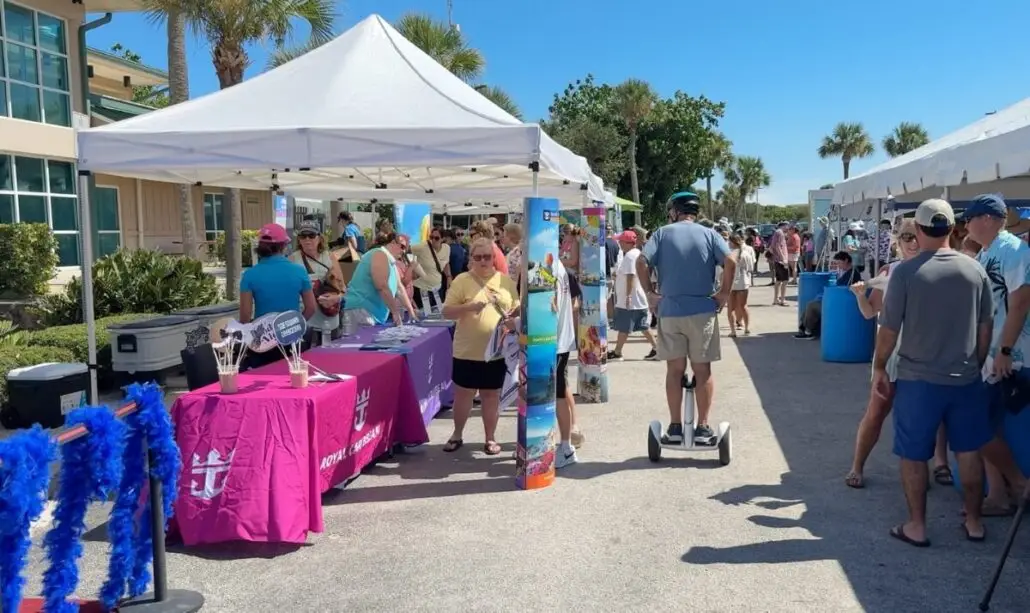 A crowd gathers at Port Canaveral for an outdoor event with vendor booths under white tents. One person rides a Segway as others browse tables with brochures and Star of the Seas inaugural sailing promotional items beneath palm trees and blue skies.