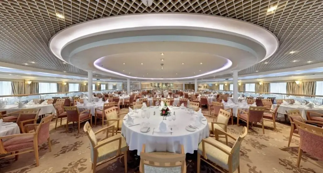 A spacious, elegant dining hall reminiscent of a cruise ship features round tables set with white tablecloths, glassware, and napkins. Wooden chairs surround the tables, and large windows line the walls under bright ceiling lights.