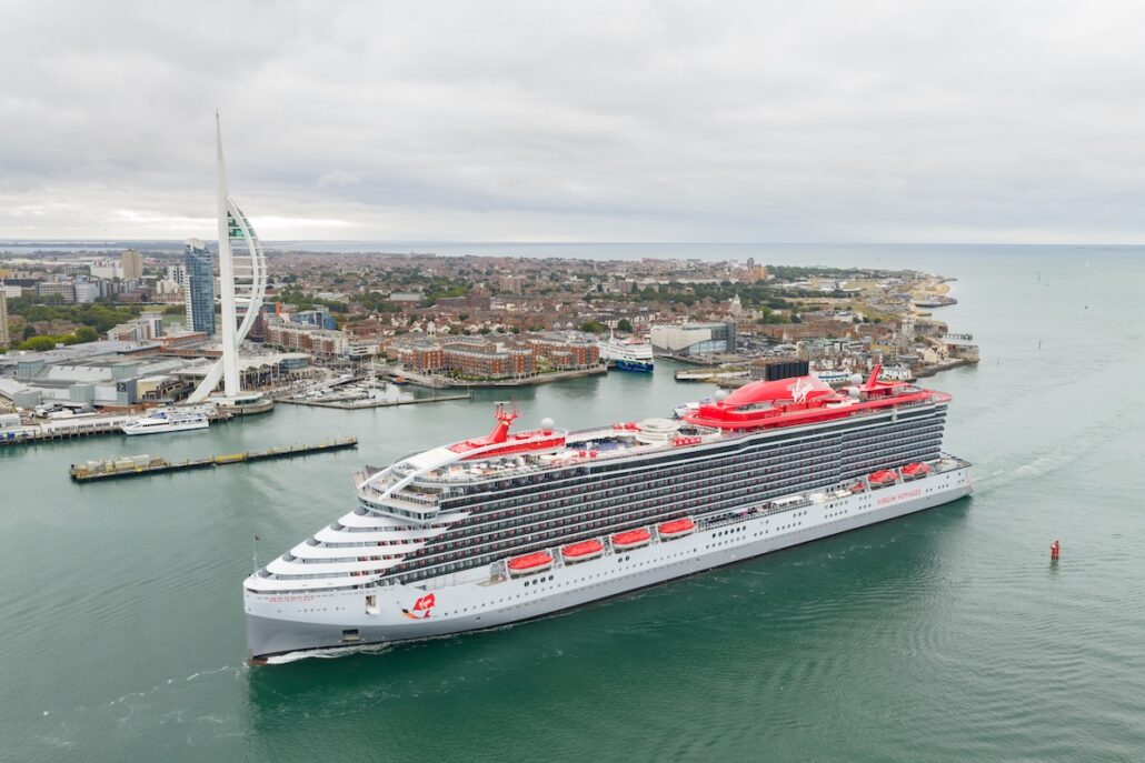 A large modern cruise ship with red and white accents sails through a harbor near a city. A tall, white, sail-shaped tower and various buildings are visible along the shoreline under a cloudy sky.