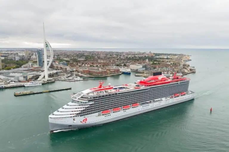 A large modern cruise ship with red and white accents sails through a harbor near a city. A tall, white, sail-shaped tower and various buildings are visible along the shoreline under a cloudy sky.