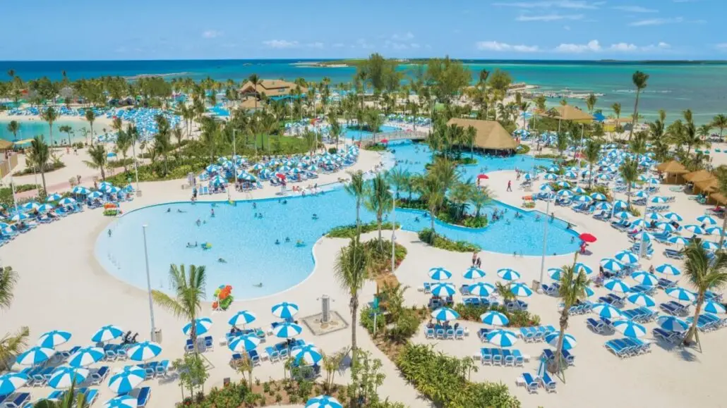 A large tropical resort with multiple pools, palm trees, many blue-and-white striped umbrellas, sun loungers, and people sunbathing or swimming—just steps from the ocean under a clear blue sky—feels as lively as a cruise terminal during peak season.