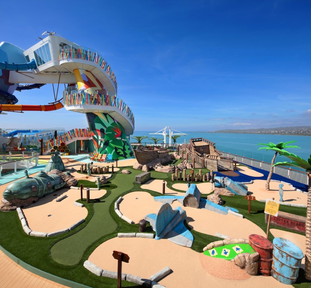 A brightly colored mini-golf course with whimsical decorations, palm trees, and a water slide sits on the deck of a cruise ship, overlooking the ocean under a clear blue sky.