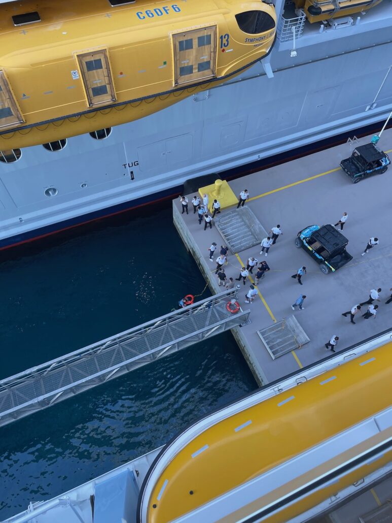 A view from above of a Royal Caribbean cruise ship docked at a pier, showing cruise passengers standing near the gangway, with yellow lifeboats attached to the ship and a small utility vehicle nearby.