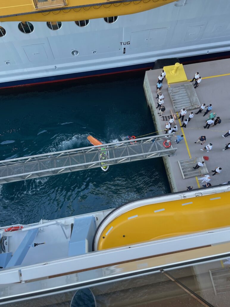 An overhead view of a docked Royal Caribbean cruise ship, a rescue boat in the water, and cruise passengers along with uniformed crew gathered on the pier near a gangway connecting the ship to CocoCay.
