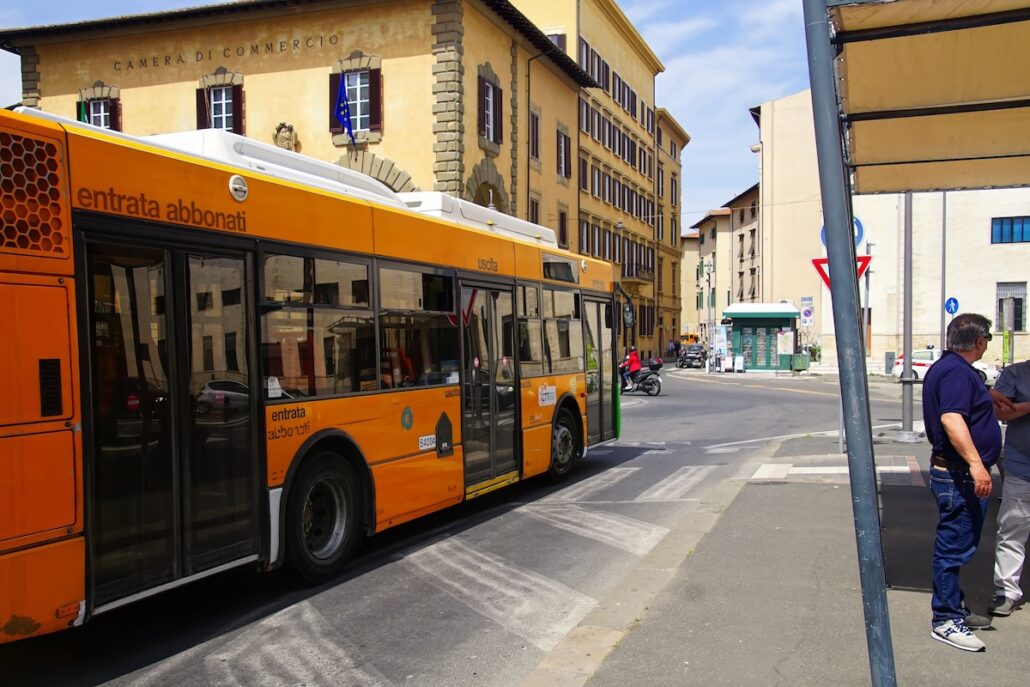 A yellow city bus stopped at a bus stop in an urban area with people standing nearby—reminiscent of the busy port where a recent port bus accident led to tragedy for an American couple. Old yellow and beige buildings stand under a partly cloudy sky.