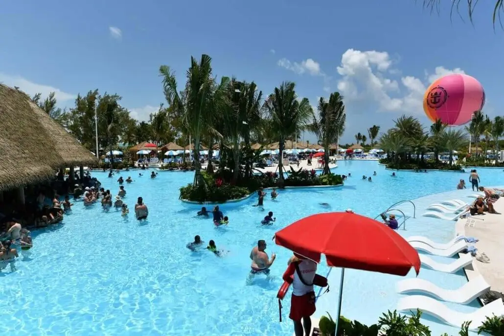 People swim and relax in a large outdoor pool on Royal Caribbean’s private island, surrounded by palm trees and lounge chairs. A lifeguard stands under a red umbrella, while a pink balloon floats in the sunny, partly cloudy sky.