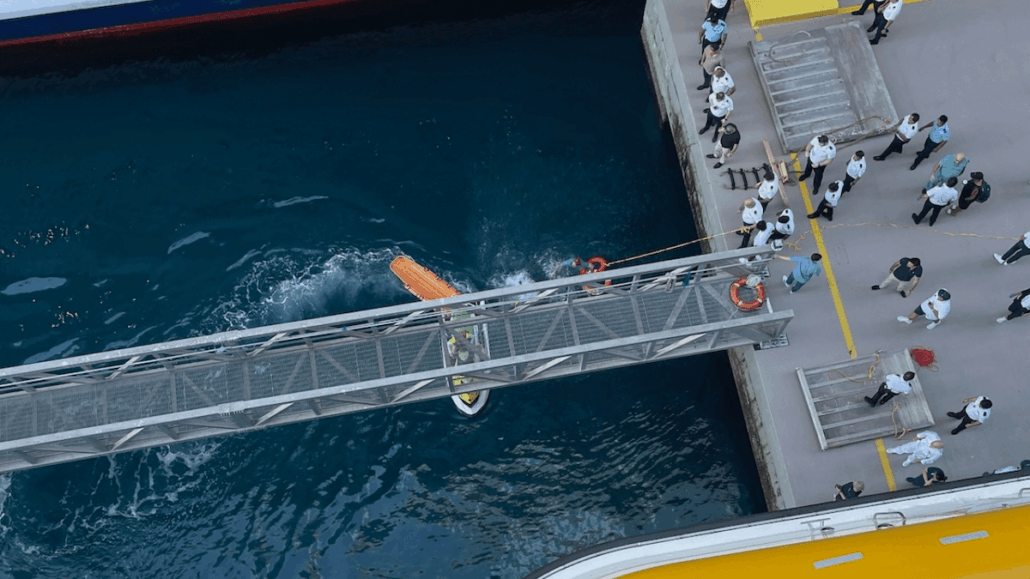 A view from above showing a lifeboat near a pier at CocoCay, with a metal gangway above the water and groups of Royal Caribbean cruise passengers standing on the dock observing the scene.