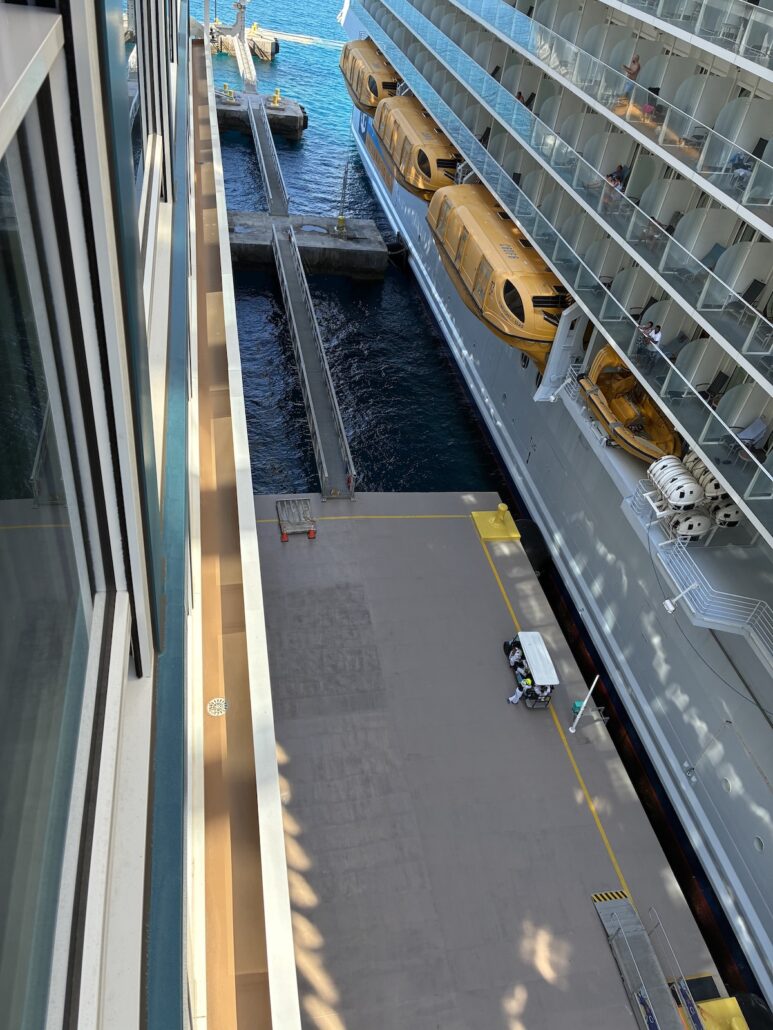 A view from above shows a Royal Caribbean cruise ship docked at the pier with yellow lifeboats in view. The mostly empty pier, possibly at CocoCay, has a cart. Blue water shimmers between the ship and dock as cruise passengers explore ashore.