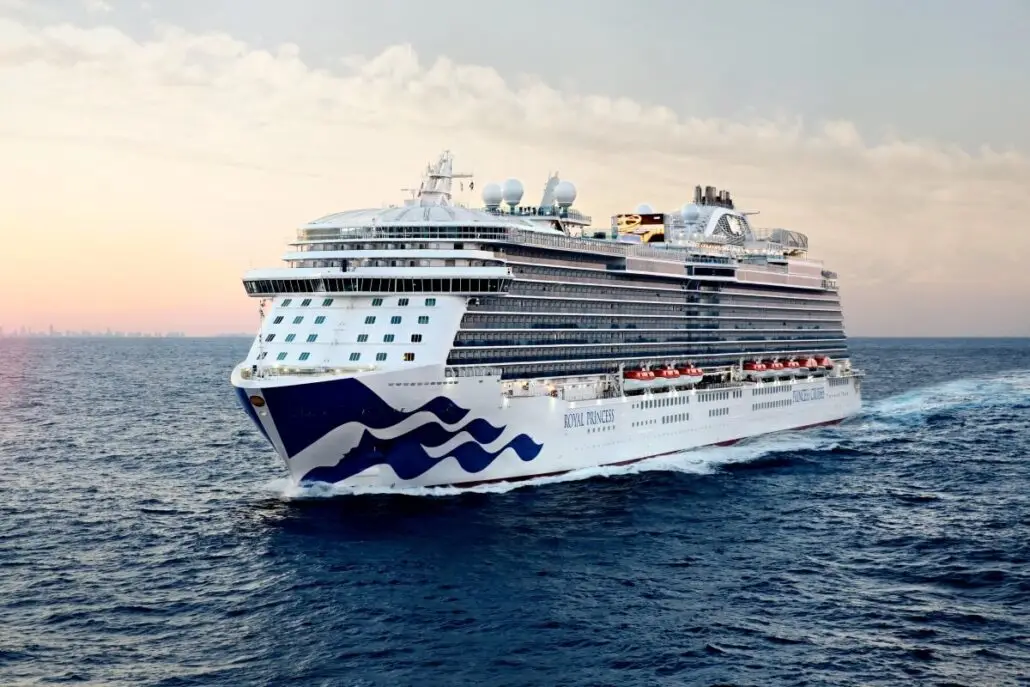 The Royal Princess, a large white cruise ship from Princess Cruises, sails across calm blue ocean water at sunset under a partly cloudy sky, with a distant city skyline hinting at exciting Asia sailings on the horizon.