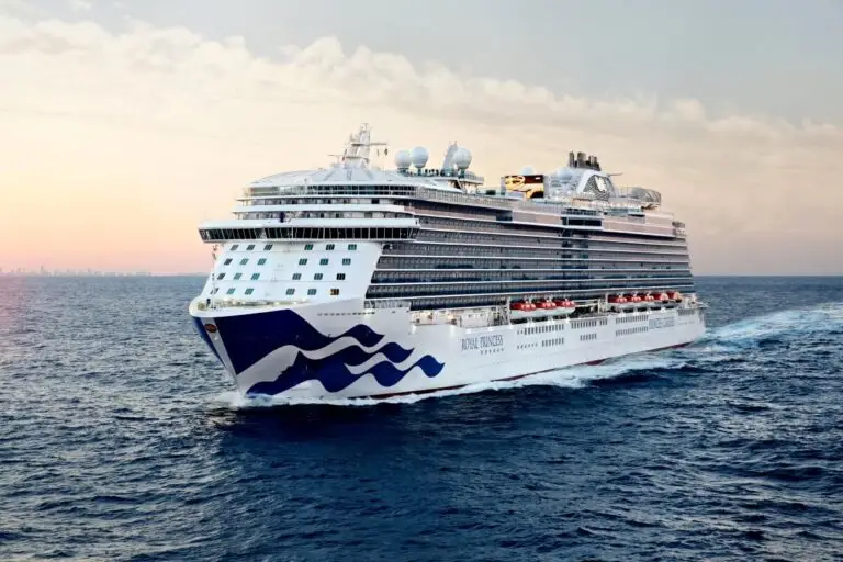The Royal Princess, a large white cruise ship from Princess Cruises, sails across calm blue ocean water at sunset under a partly cloudy sky, with a distant city skyline hinting at exciting Asia sailings on the horizon.