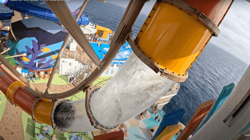 A clear tube water slide with a sharp turn descends from an upper deck towards a colorful pool area on a cruise ship, with the ocean visible in the background—perfect fun for families where every child enjoys safely.