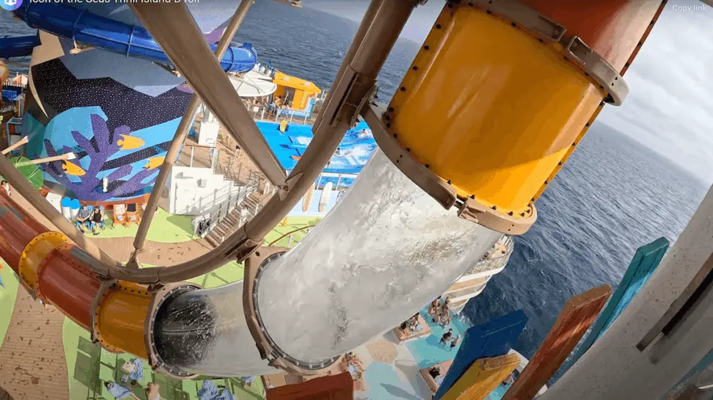 A clear tube water slide with a sharp turn descends from an upper deck towards a colorful pool area on a cruise ship, with the ocean visible in the background—perfect fun for families where every child enjoys safely.