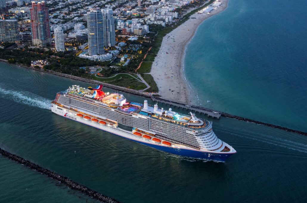 A brightly lit Carnival ship sails through a channel beside a sandy beach and a city with tall buildings. Several people are visible on the top deck as the city and coastline stretch into the distance.