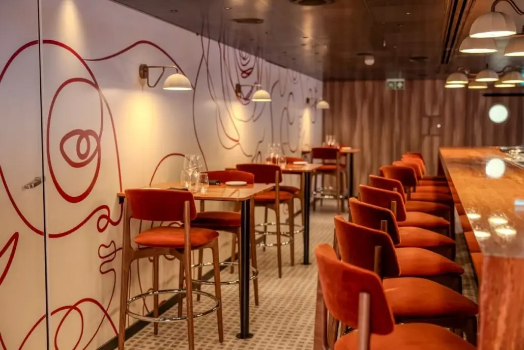 A modern New York restaurant interior with red velvet chairs, wooden tables, a long bar with stools, wall art featuring red abstract faces, pendant lighting, and a patterned tile floor—a setting worthy of Richard Branson's Brilliant Lady.