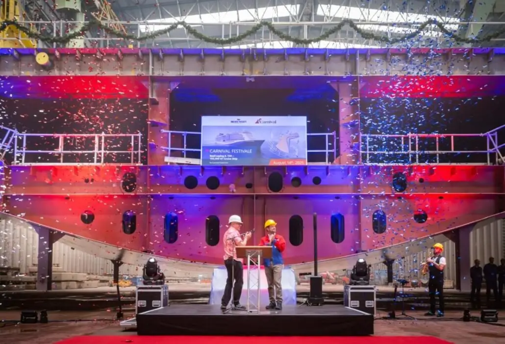 Two people in hard hats stand at a podium on a red carpet before a massive LNG flagship under construction, with confetti falling and a Carnival Festivale banner overhead. Other cruise line workers and equipment are visible in the background.