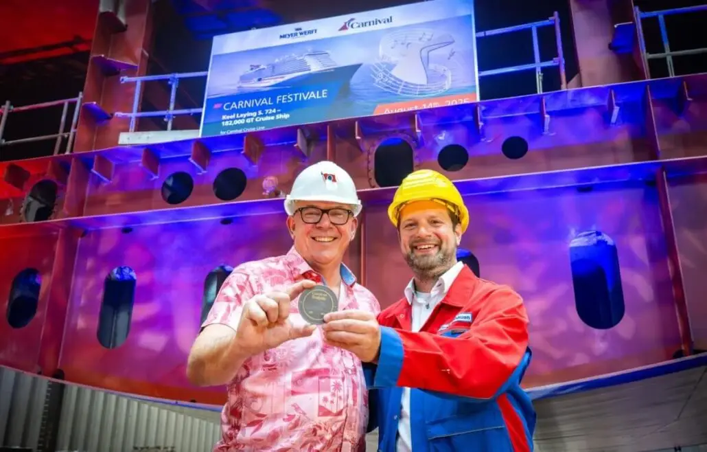 Two men in hard hats and safety gear smile and hold a commemorative coin in front of a large ship section. A Carnival Festivale cruise line launch event banner is displayed above them, celebrating the LNG flagship's debut.