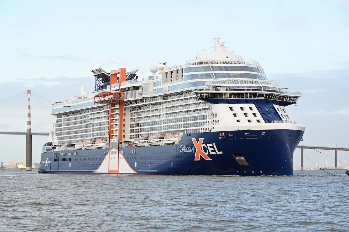 A large blue and white cruise ship labeled "Celebrity Xcel" is sailing on calm water near a bridge. Undergoing sea trials ahead of its November launch, the ship has multiple decks, orange lifeboats, and a distinctive orange structure on the side.