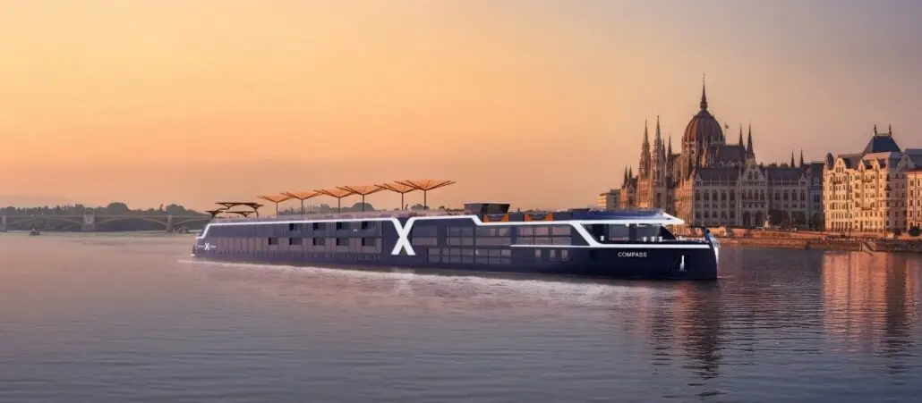 A modern river cruise ship sails on a calm river at sunset, passing historic European buildings, including a large, ornate parliament structure—an idyllic scene for the 2027 river cruise season and upcoming cruise sales.
