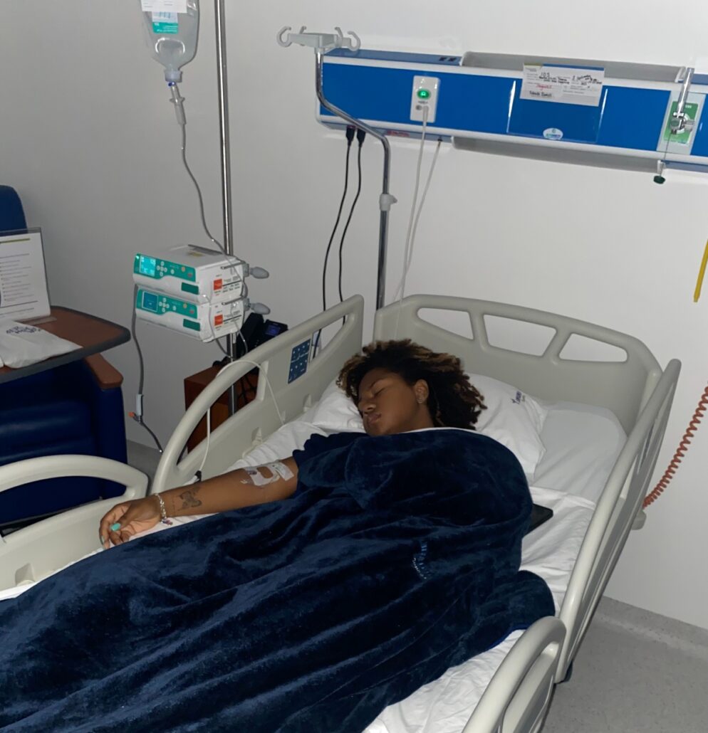 A Texas family member lies in a hospital bed with an IV, covered by a dark blanket. Medical equipment hangs on the wall and a chair sits nearby, while dim lighting hints at concern over an impending medical bill.