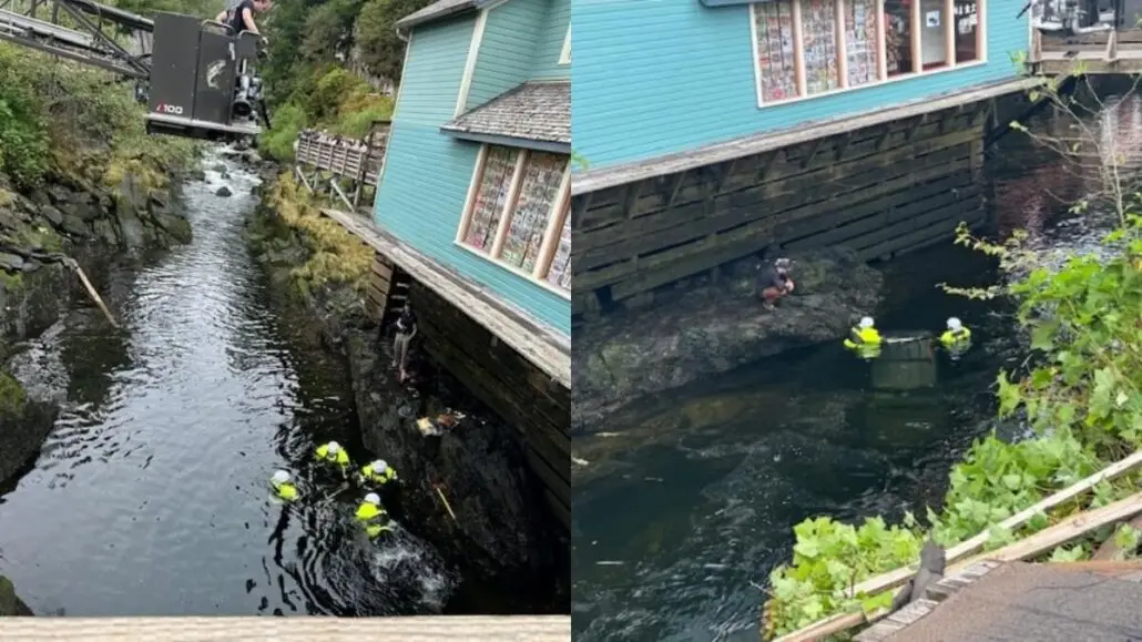 Several people in yellow safety gear work in a narrow stream beside a blue-sided wooden building in Ketchikan, Alaska. One person climbs a ladder attached to the building, while others are in the water near the rocky bank after a car accident.