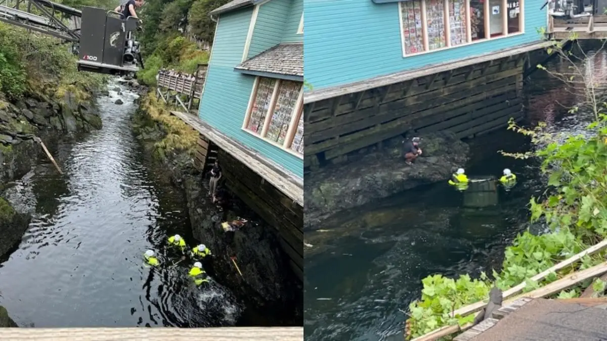 Several people in yellow safety gear work in a narrow stream beside a blue-sided wooden building in Ketchikan, Alaska. One person climbs a ladder attached to the building, while others are in the water near the rocky bank after a car accident.