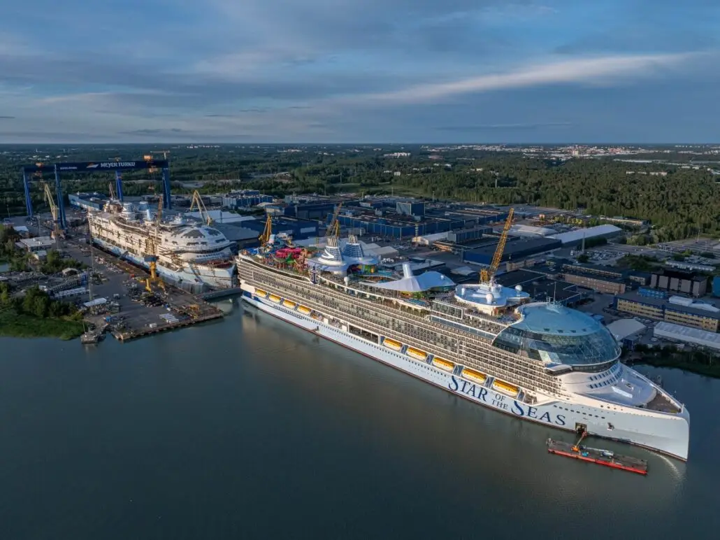 A large cruise ship named "Star of the Seas," part of Royal Caribbean, is docked at Meyer Turku Shipyard beside another vessel. Several cranes surround the ships, with industrial buildings and a forested area in the background, highlighting modern shipbuilding.