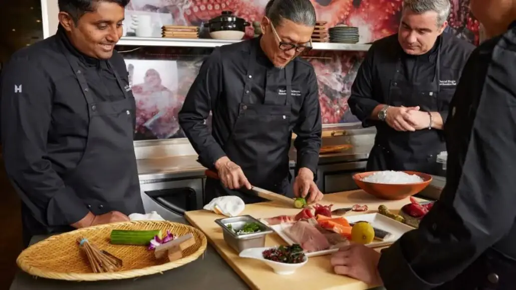 Three chefs in black uniforms, including Iron Chef Morimoto, stand around a kitchen counter as one of them slices fish. Various ingredients and garnishes are arranged on the counter, evoking the culinary excellence of Holland America 2026 Grand Voyages.