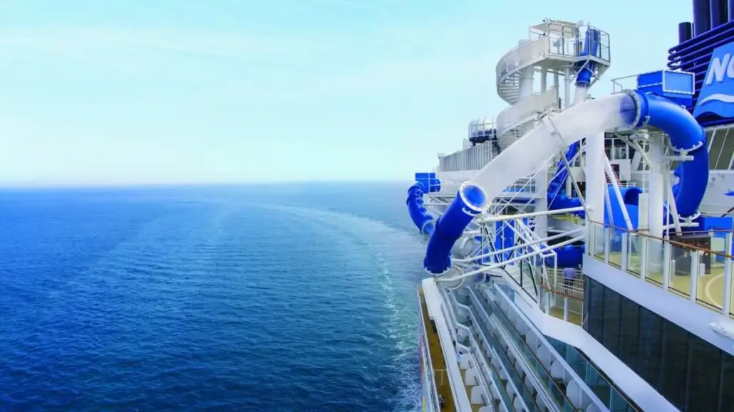 A large cruise ship is sailing on the calm ocean, leaving a trail behind. Clear and blue enclosed water slides extend over the side, offering cruise passengers an exciting view. The sky is clear, making it perfect for a viral TikTok moment.