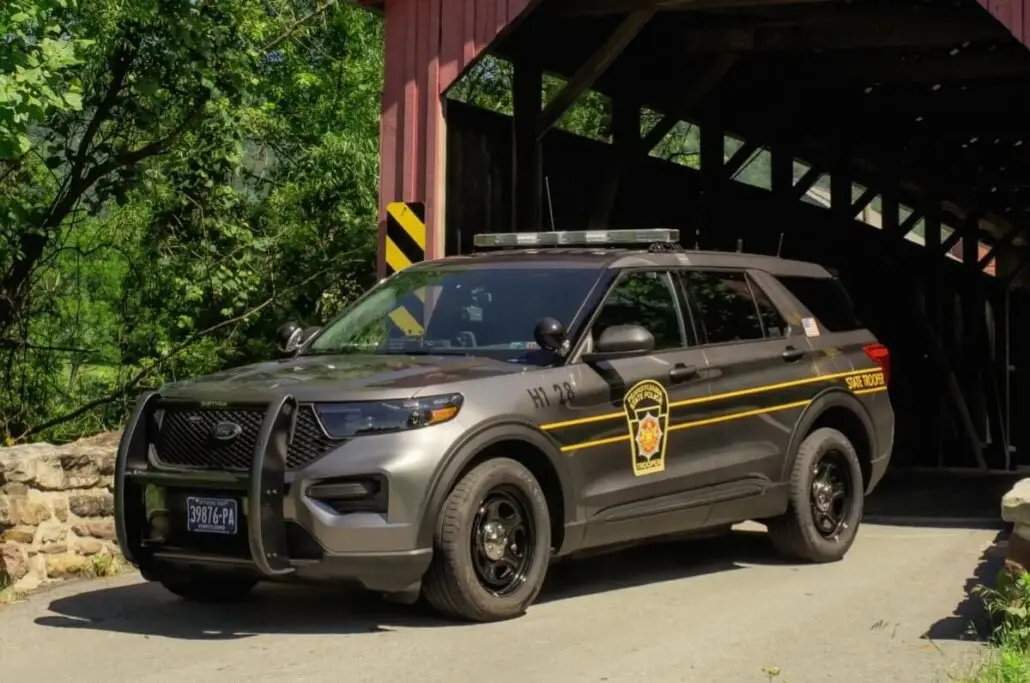 A black and gray state police SUV is parked on a road in front of a red covered bridge, surrounded by greenery. The vehicle, with visible police markings and emergency lights, appears to be responding to an incident involving an injured relative.