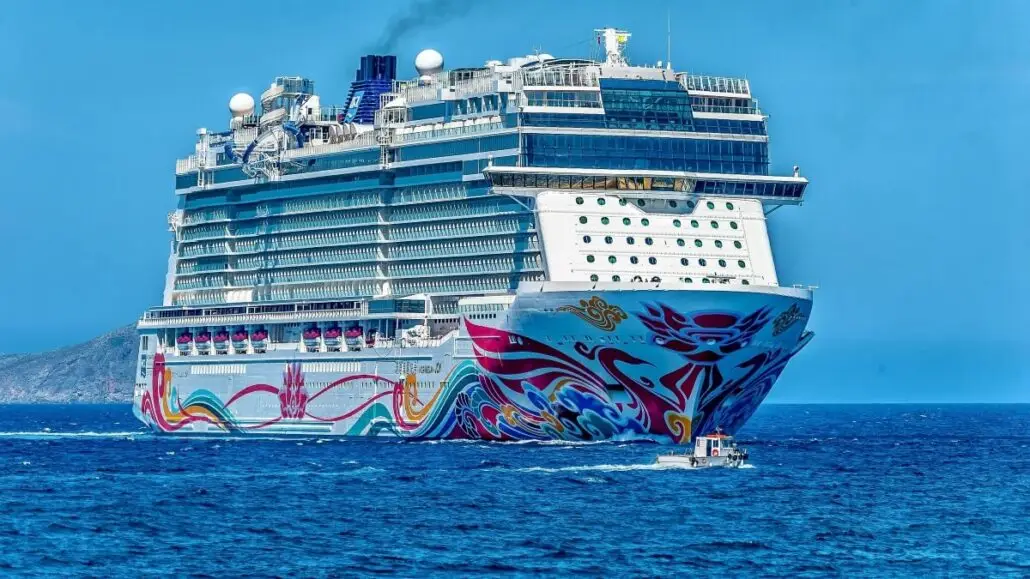 A large, modern cruise ship with colorful designs on its hull sails on blue Caribbean waters under a clear sky, passing a small boat with distant islands and tropical ports visible in the background.