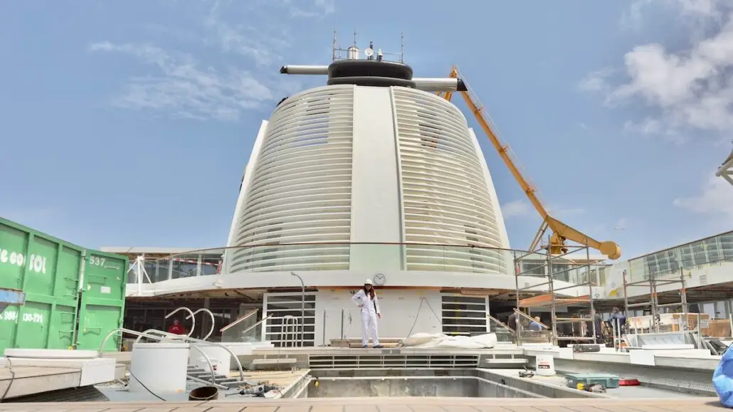 A worker in a white jumpsuit stands in front of a large, modern World Residential Yacht structure at dry dock; a yellow crane, construction materials, and containers are visible under a partly cloudy sky.
